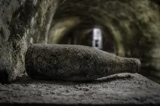 Dusty champagne bottle resting in a dimly lit chalk cellar with cool, gray-green hues and arched stone ceiling.