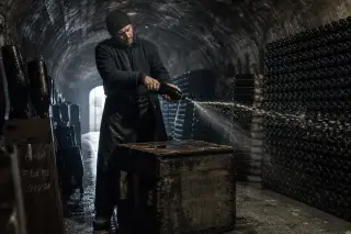 Vigneron degorging a champagne bottle by hand in a cold, dimly lit cellar lined with rows of unlabeled bottles and chalk-streaked stone walls.