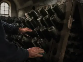 Riddler's hands turning champagne bottles on a wooden pupitre in a dim, arched cellar with rows of dusty bottles, cold natural light highlighting textured surfaces.