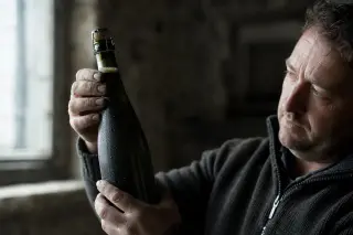 Vigneron holds up a champagne bottle to the light in a cold cellar, focusing on the bubbles and cork, hands weathered from vineyard work, soft grey-green tones throughout.